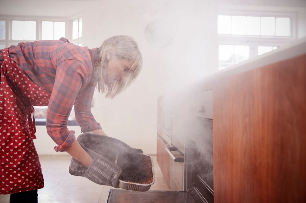 A woman in an apron cooks in an oven, surrounded by signs of smoke damage and soot residue in the kitchen.