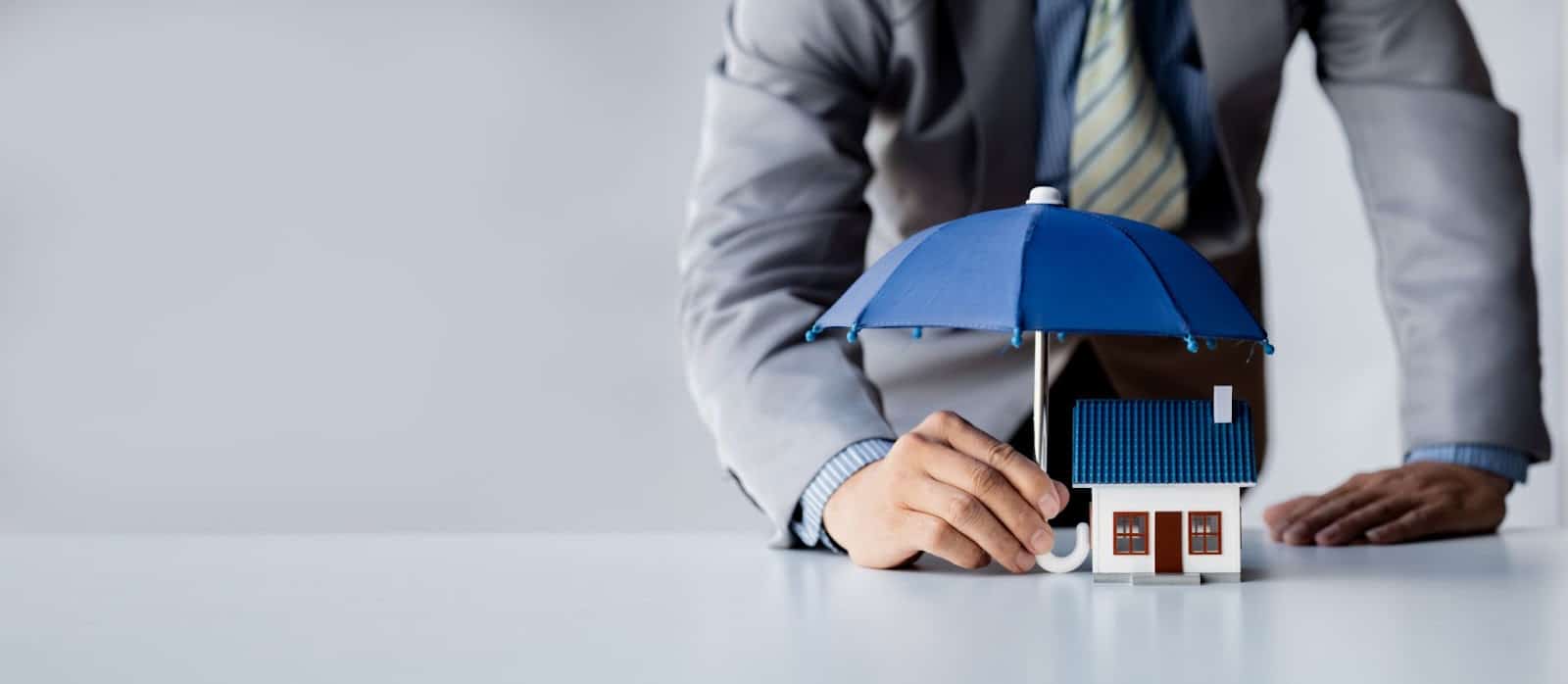 Businessman protects a model house with an umbrella, symbolizing storm damage insurance and disaster preparedness.